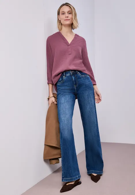 Frau trägt rosa Pullover, blaue Jeans mit weitem Bein, schwarze Pumps, hält braune Jacke.